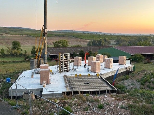 Chantier de construction avec dalle en béton, briques, grue et terres agricoles en arrière-plan.