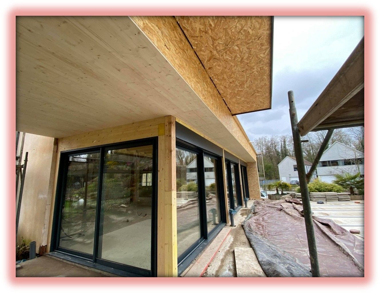 Extérieur d'un bâtiment en construction, avec de grandes baies vitrées, des poutres en bois et des échafaudages.