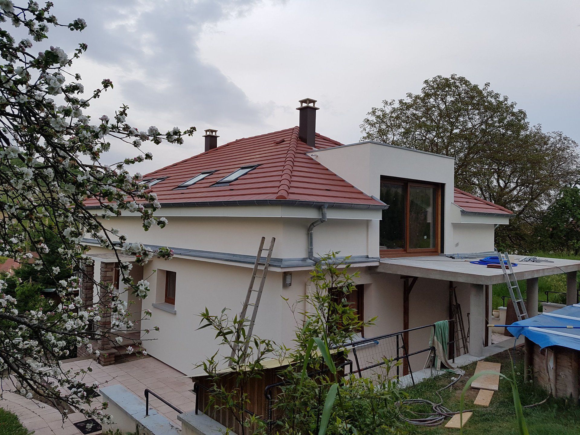 Maison moderne à deux étages avec toit de tuiles rouges, en construction, entourée d'arbres, ciel nuageux.
