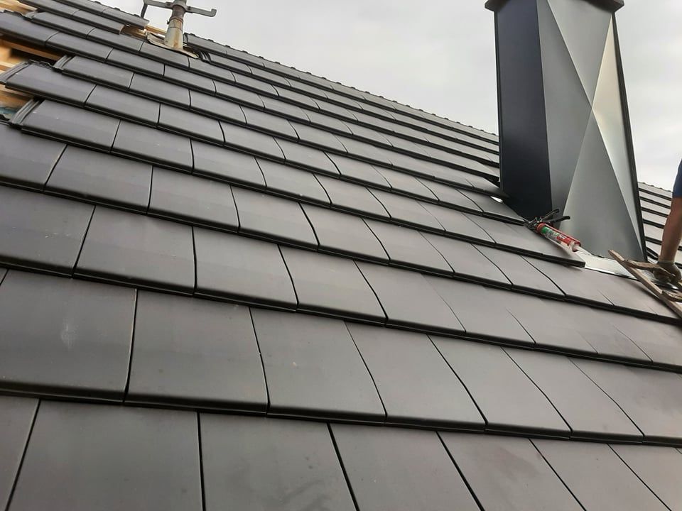 Pose de tuiles gris foncé, cheminée noire visible.