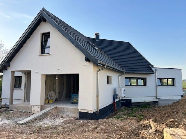 Maison blanche en construction, toit sombre, garage ouvert, encadrements de fenêtres, climatiseur, aménagement extérieur.