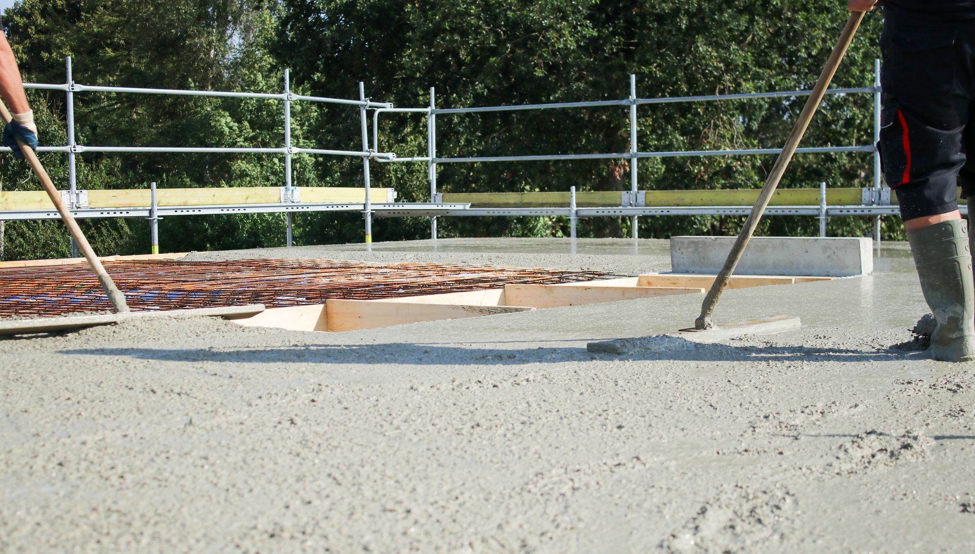 Des ouvriers lissent du béton frais sur un chantier de construction à l'aide d'outils à long manche, devant un échafaudage.