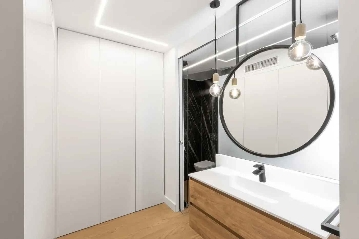 Baño moderno con tocador blanco, espejo redondo, lámparas colgantes y detalles de madera.