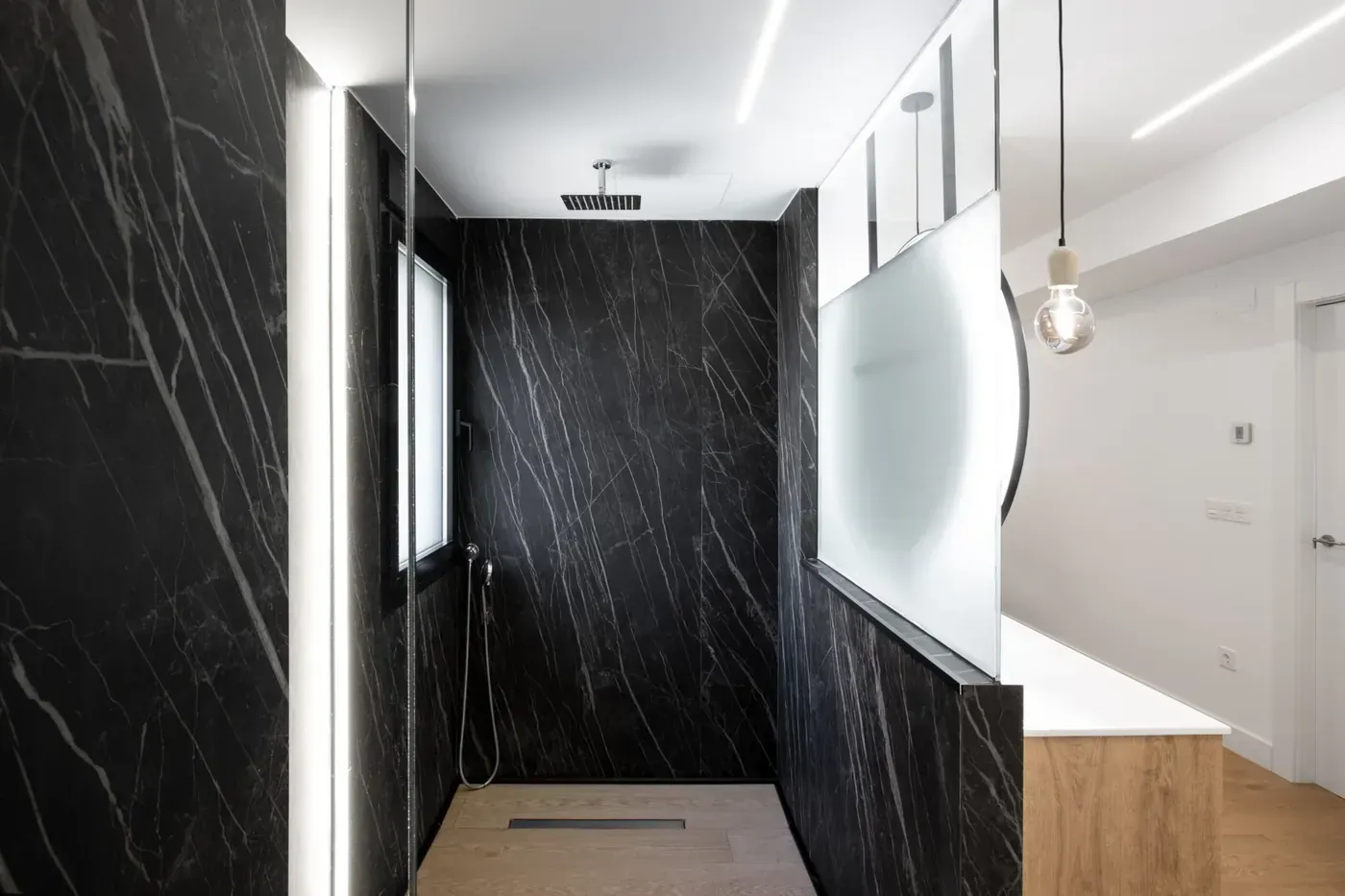 Pasillo moderno con paredes de mármol negro, techo blanco y mampara de vidrio esmerilado junto a suelo de madera.