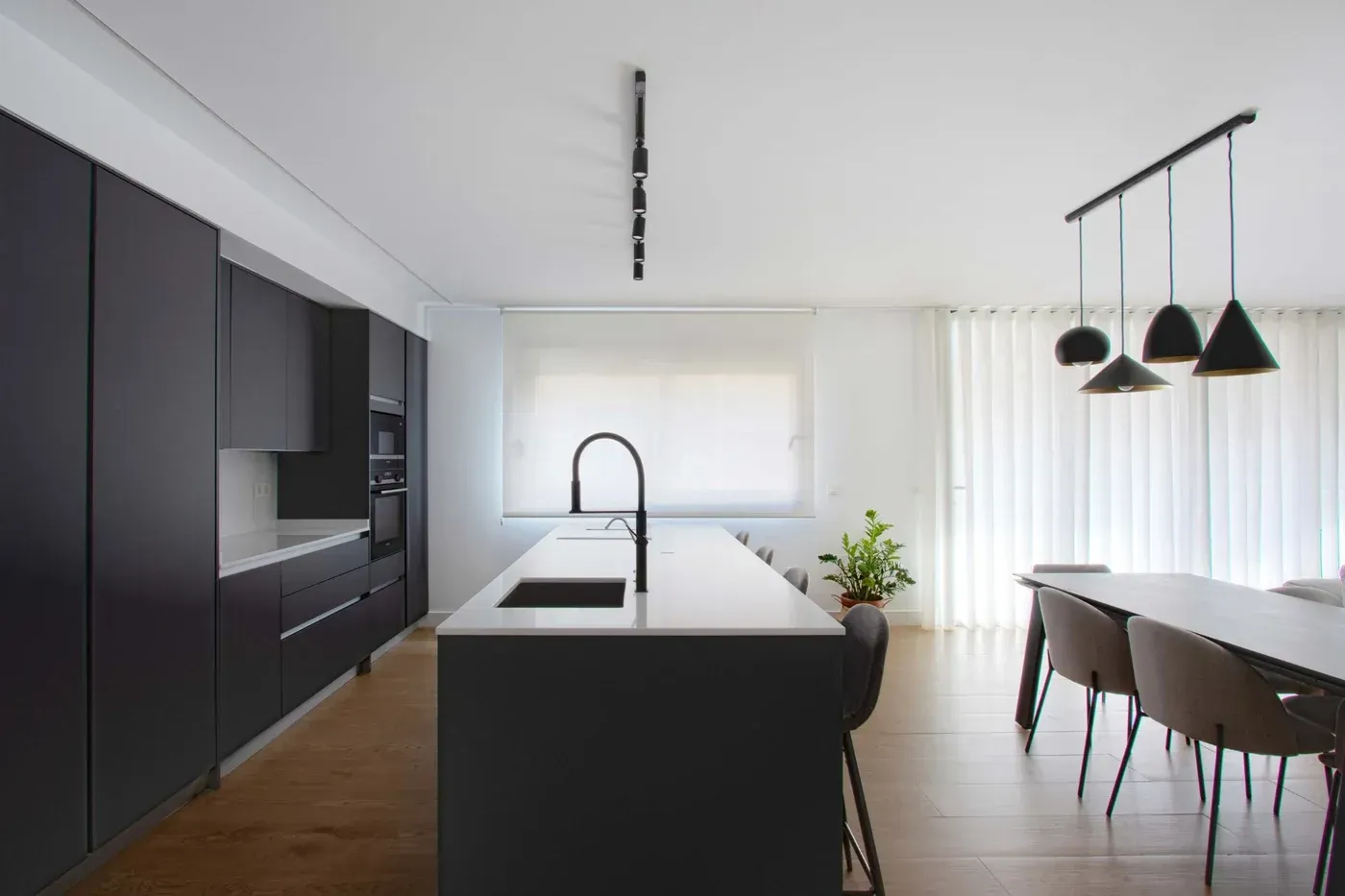 Cocina moderna en blanco y negro con isla, lámparas colgantes, mesa de comedor y gran ventanal.