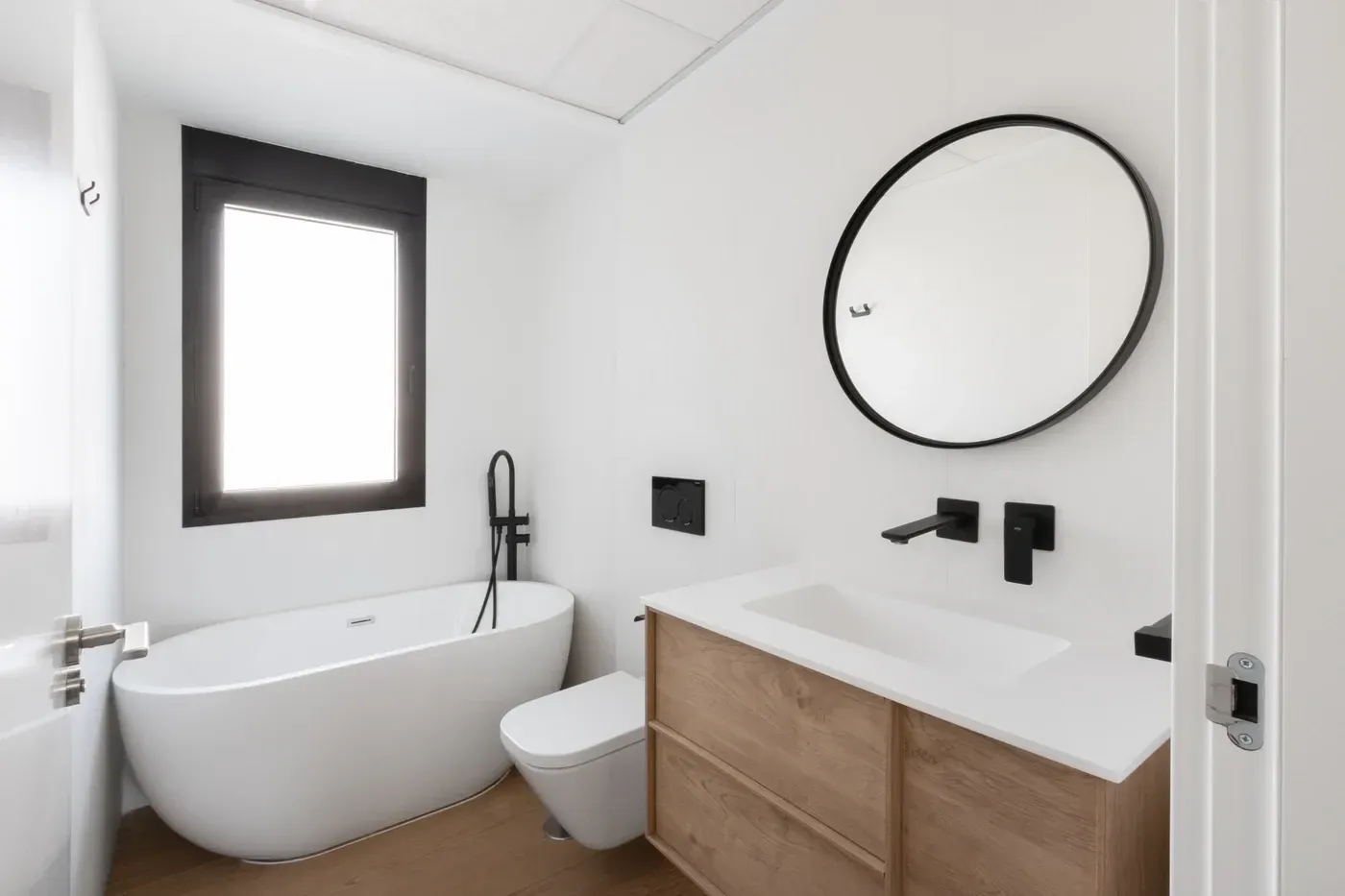 Baño moderno con bañera independiente, tocador de madera, espejo redondo y grifería negra.