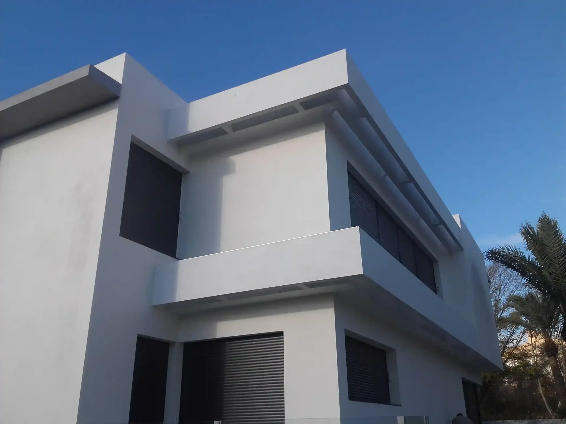 Moderna casa blanca de dos plantas con ventanas oscuras bajo un cielo azul.