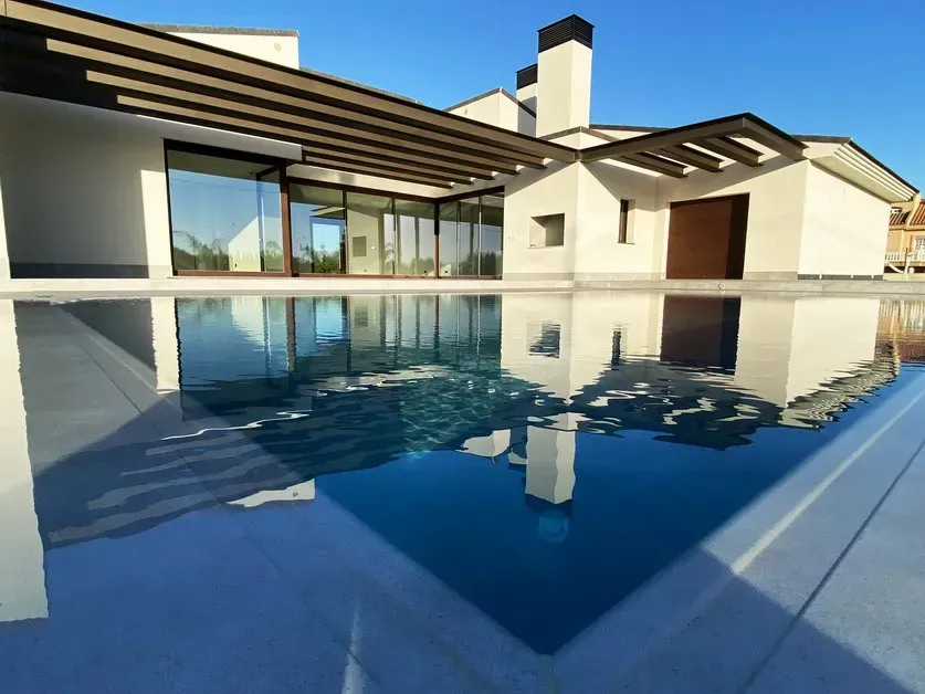 Casa moderna blanca con grandes ventanales que se reflejan en una tranquila piscina azul.