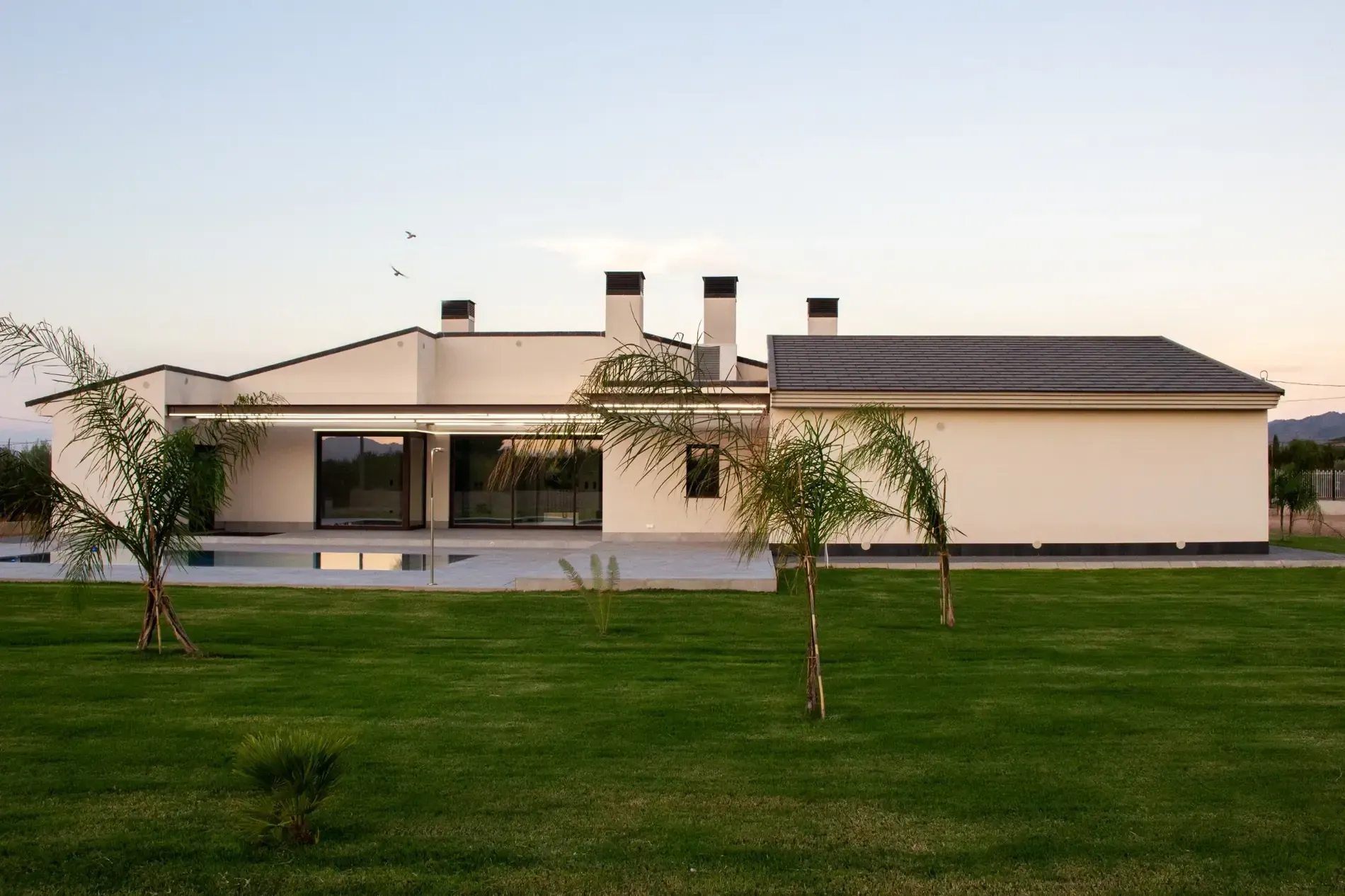 Casa blanca de una sola planta con techo oscuro, porche delantero y césped verde al atardecer.