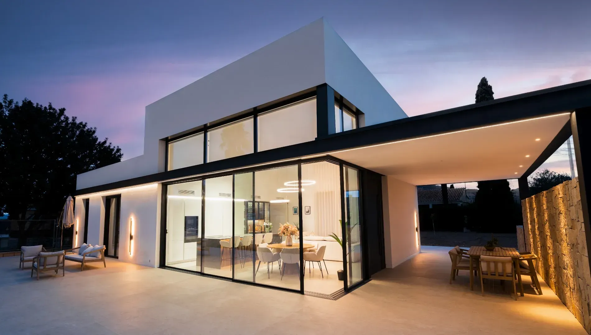 Moderna casa blanca de dos plantas con grandes paredes de cristal y un patio cubierto para disfrutar del atardecer.