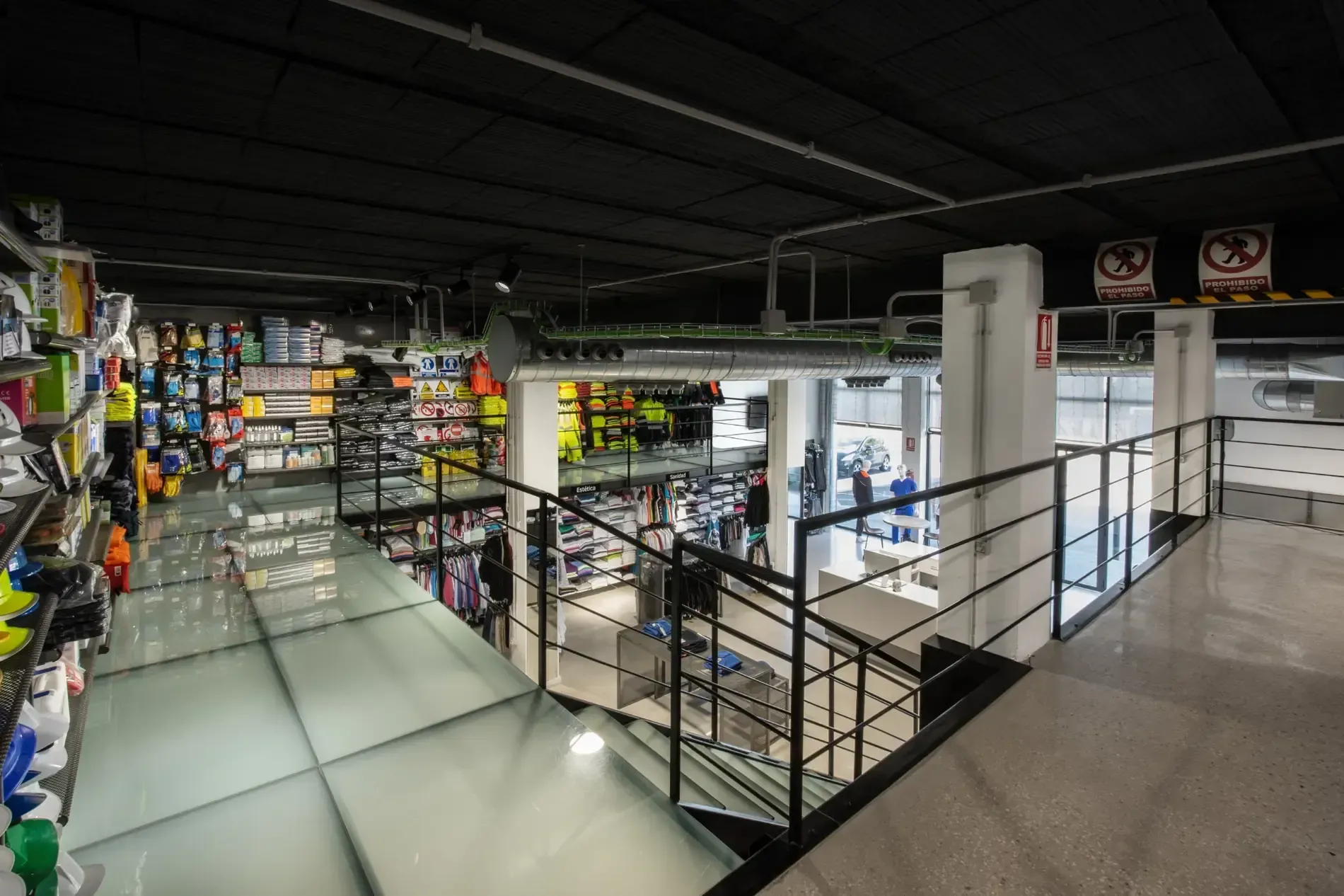 Interior de una tienda moderna con suelos de cristal, barandillas negras y estanterías con productos coloridos.