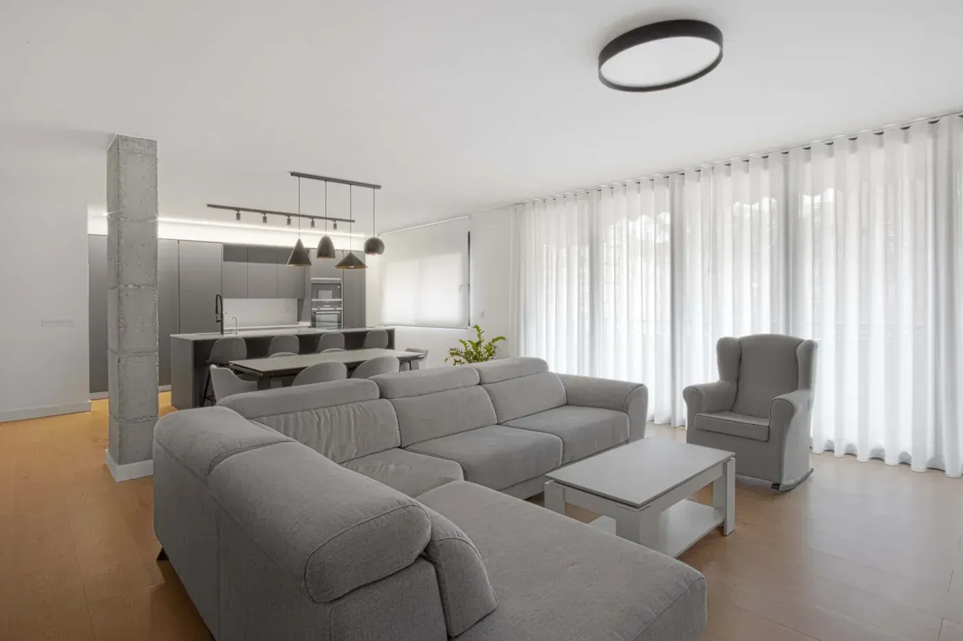 Luminoso salón moderno con sofá modular gris, zona de comedor y cortinas transparentes del suelo al techo.