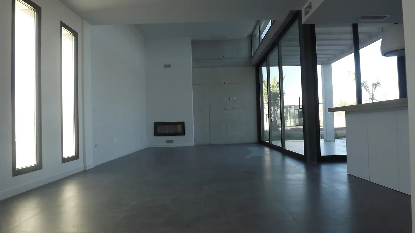 Habitación moderna vacía con suelo gris, paredes blancas, ventanas estrechas y puertas de cristal que dan a un patio.