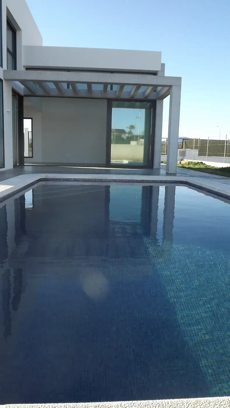 Piscina moderna de villa con agua azul y una pérgola blanca con vistas a un paisaje luminoso.