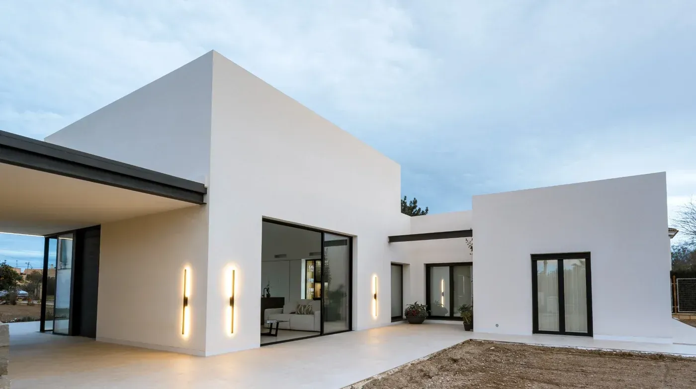 Casa moderna de estuco blanco con grandes ventanales y entrada cubierta al anochecer.