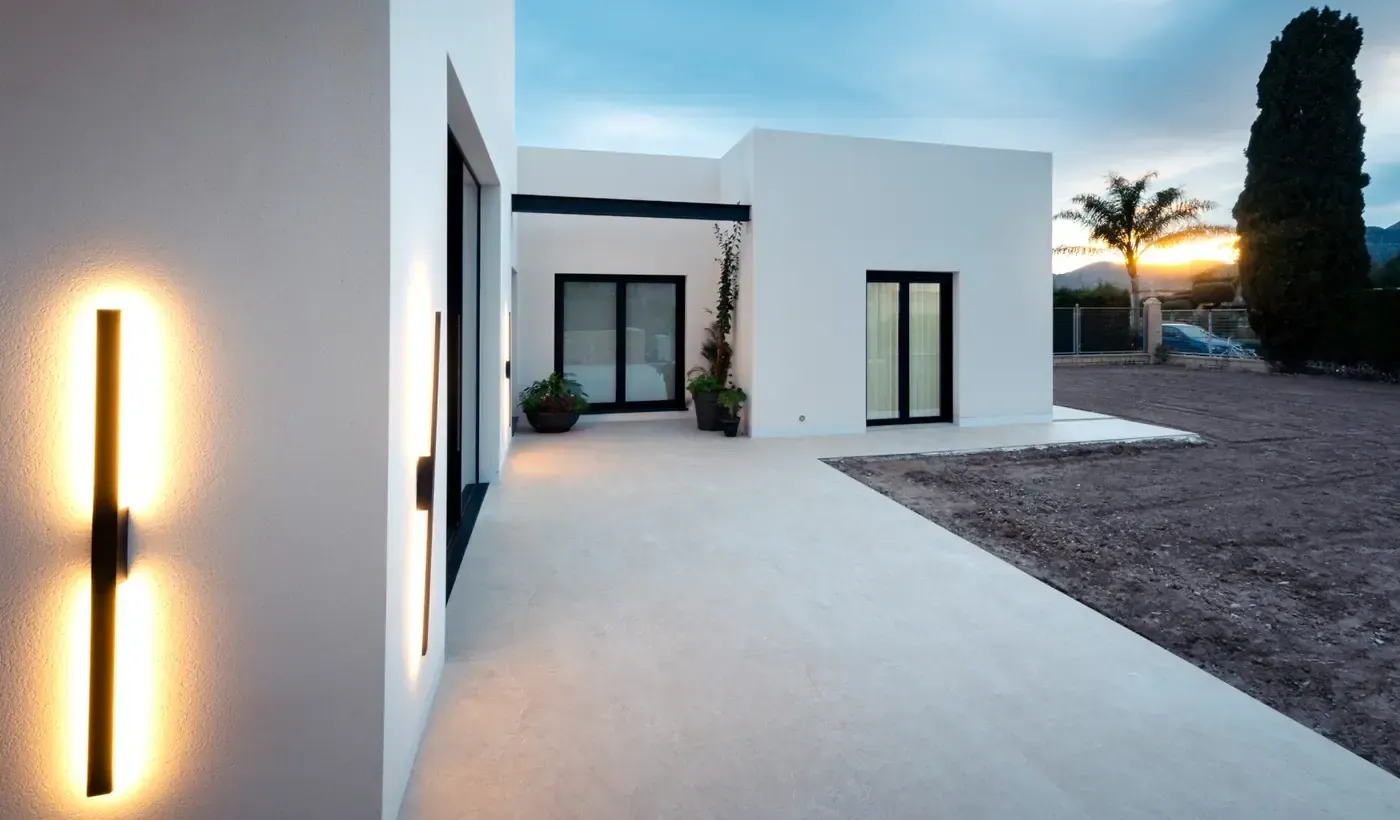 Exterior de una casa moderna blanca al atardecer con luces de pared encendidas y un patio minimalista.
