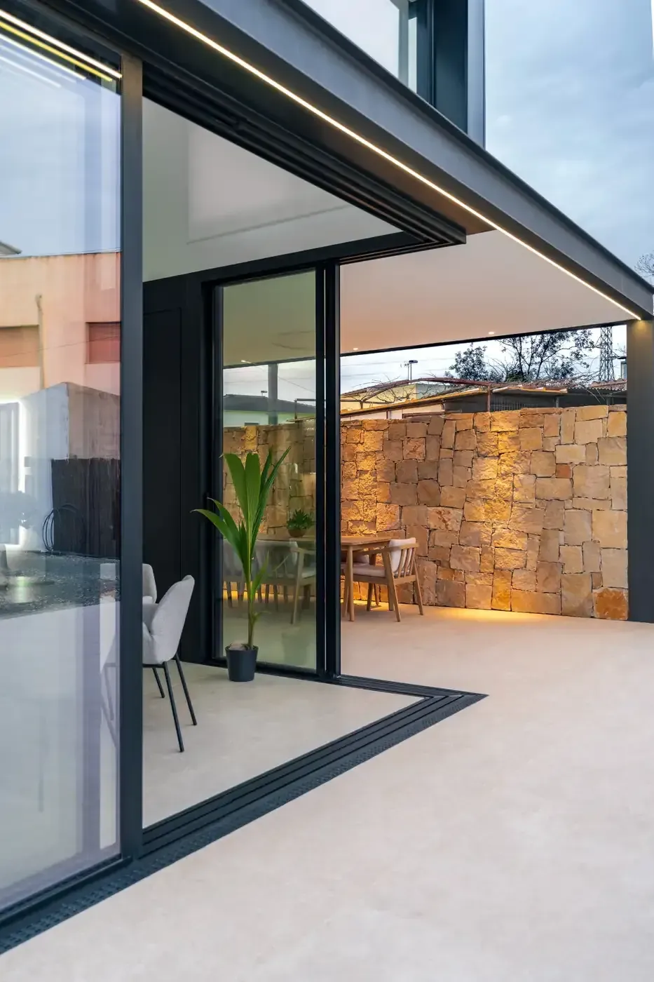 Patio moderno junto a una casa con paredes de cristal, con muro de piedra e iluminación exterior cálida al anochecer.