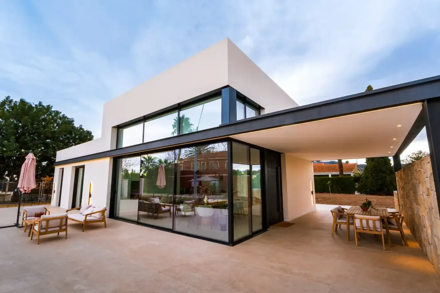 Casa moderna con grandes paredes de cristal y un patio cubierto con zona de estar al atardecer.