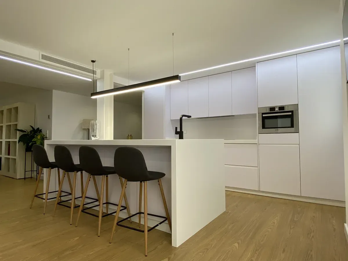 Cocina moderna blanca con una gran isla, taburetes de bar y electrodomésticos.