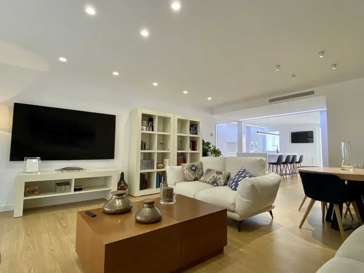 Luminoso salón moderno con sofás blancos, mesa de centro de madera, televisor de pared, estanterías y zona de comedor.