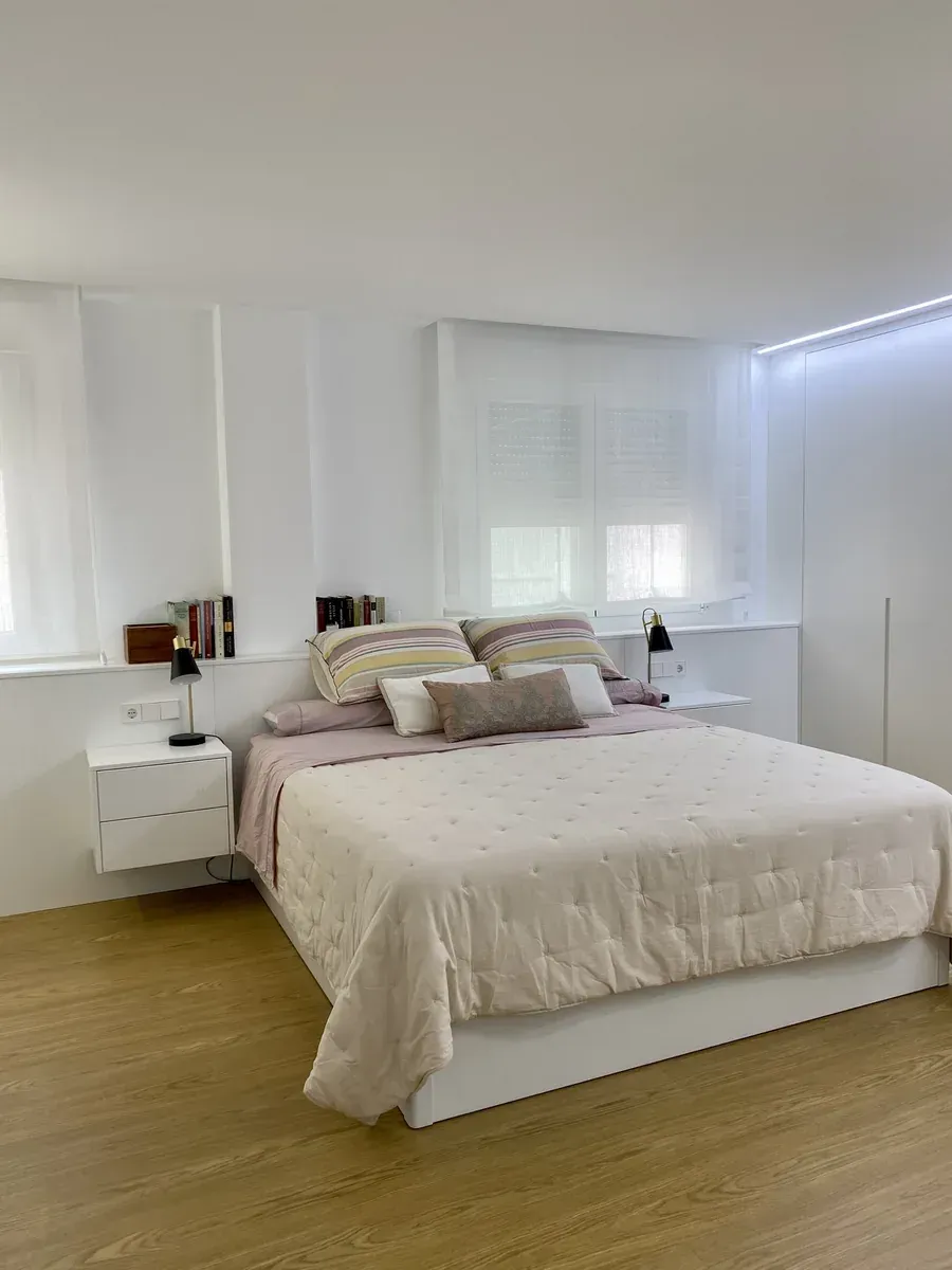 Dormitorio blanco luminoso con una cama bien hecha, mesitas de noche y suelo de madera.