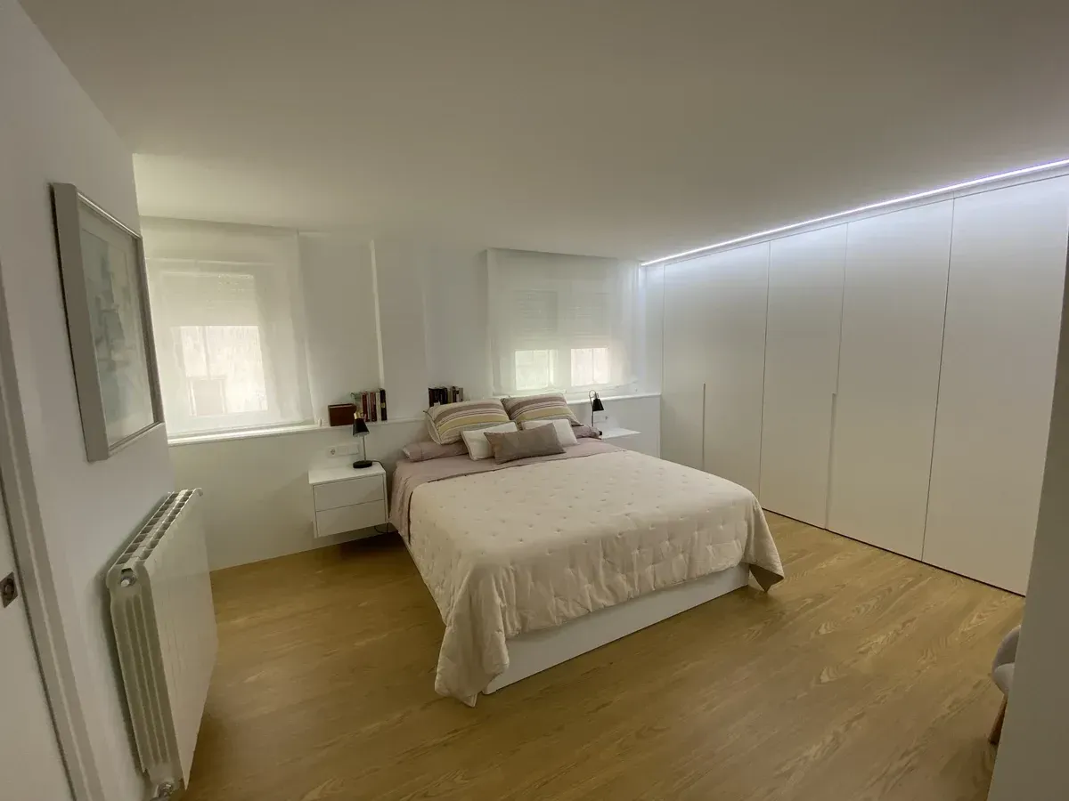 Habitación luminosa y minimalista con cama, paredes blancas, suelo de madera y puertas de armario iluminadas con LED.