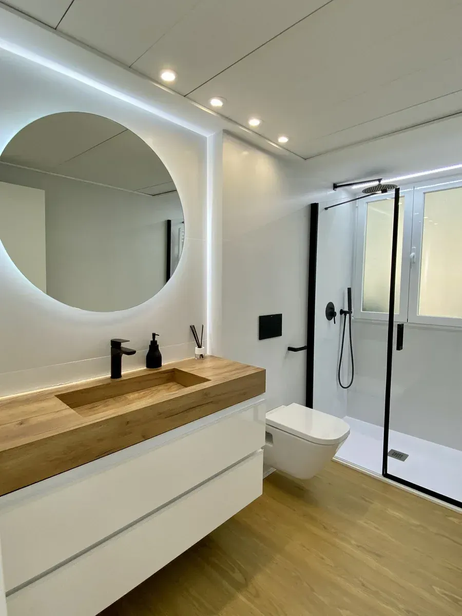 Baño moderno blanco con tocador de madera, espejo redondo y mampara de ducha de cristal.