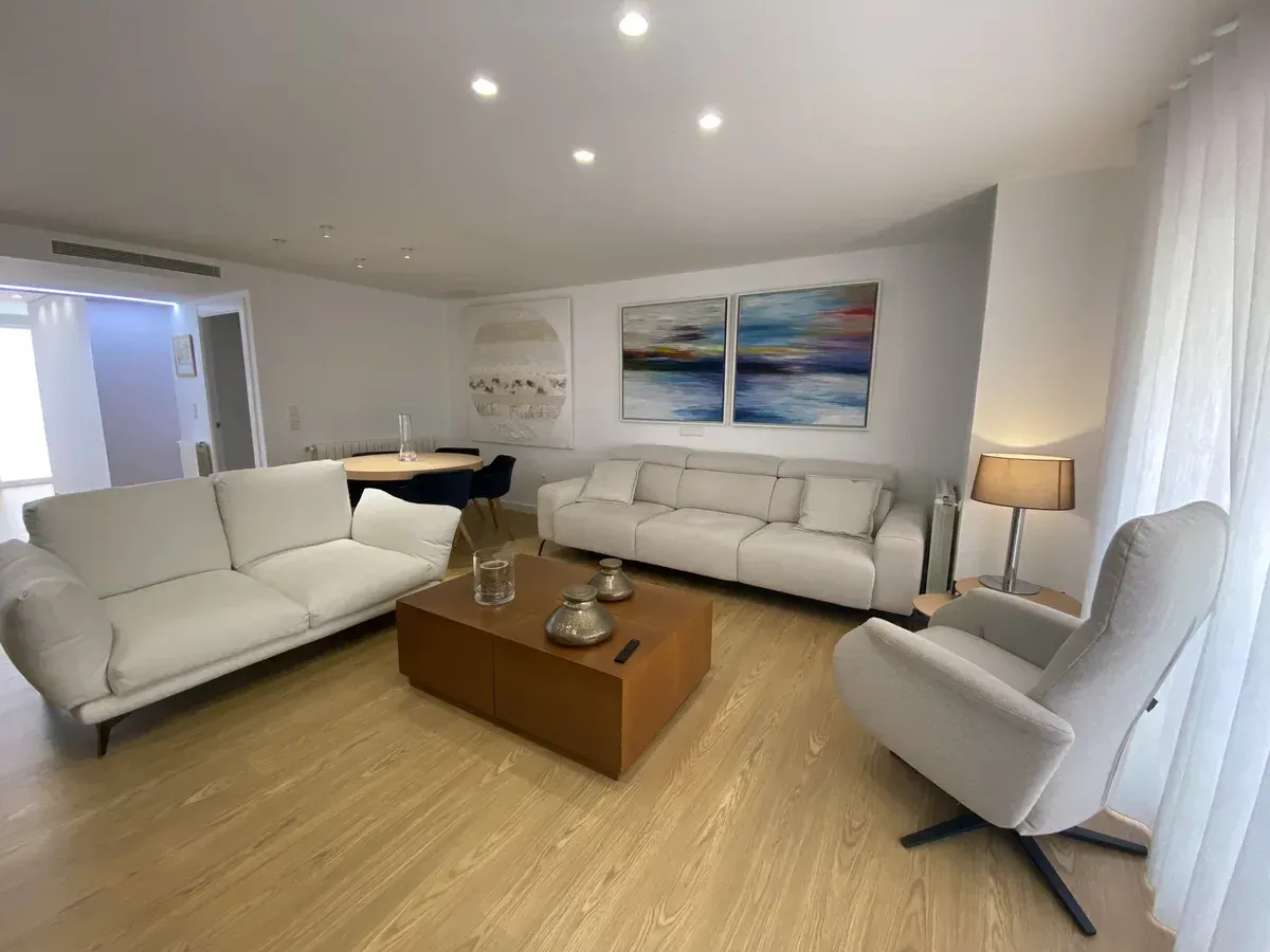 Sala de estar moderna y luminosa con sofás blancos, mesa de centro de madera, lámpara de pie y cuadro con motivos marinos.