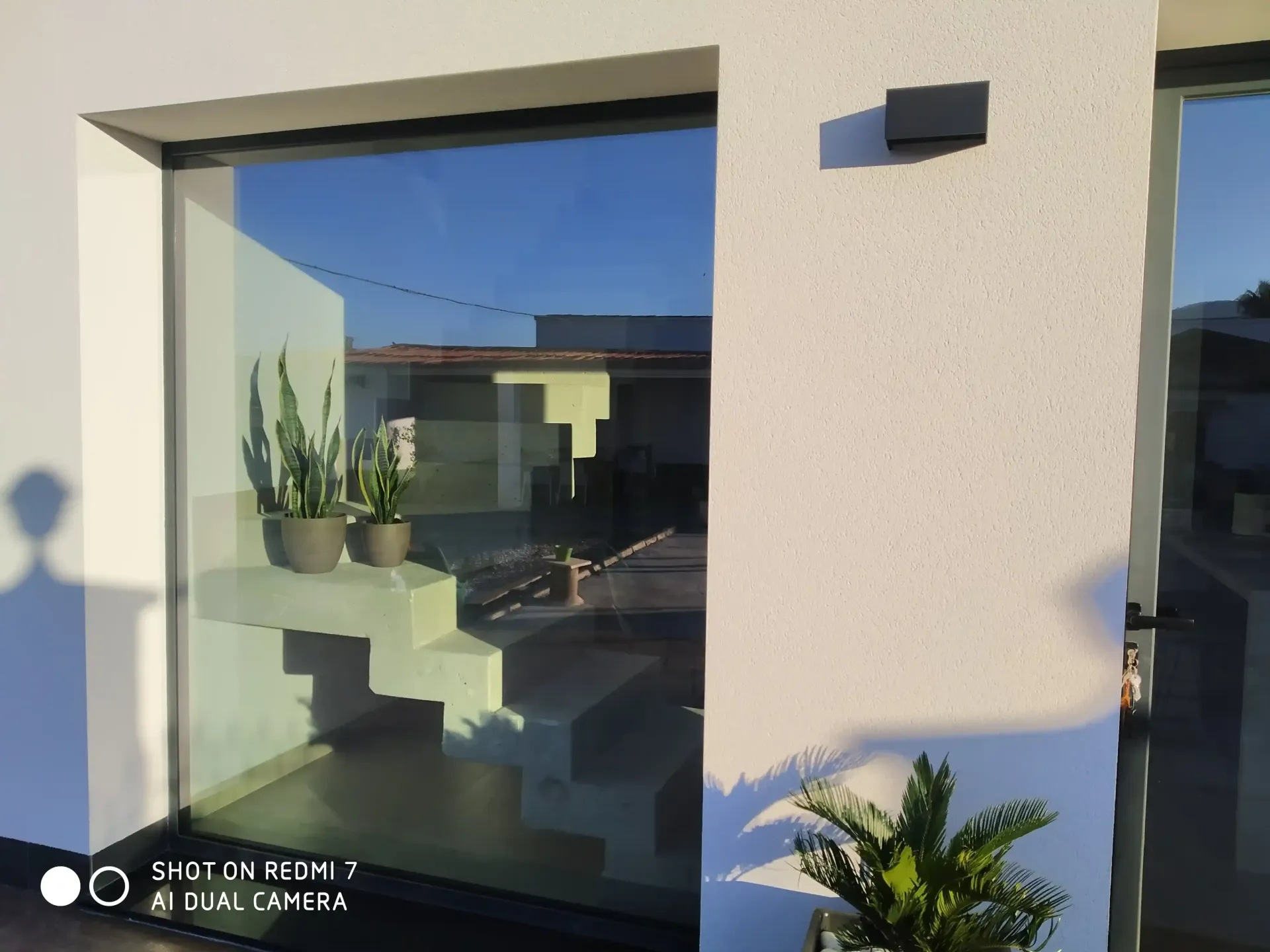 Exterior de una casa moderna con una gran ventana, plantas en macetas y una pared de estuco iluminada por el sol.