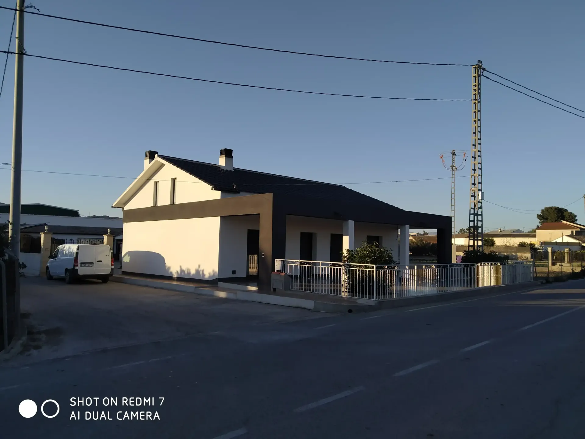 Una pequeña casa blanca en una esquina al anochecer, con postes de luz y una furgoneta aparcada cerca.