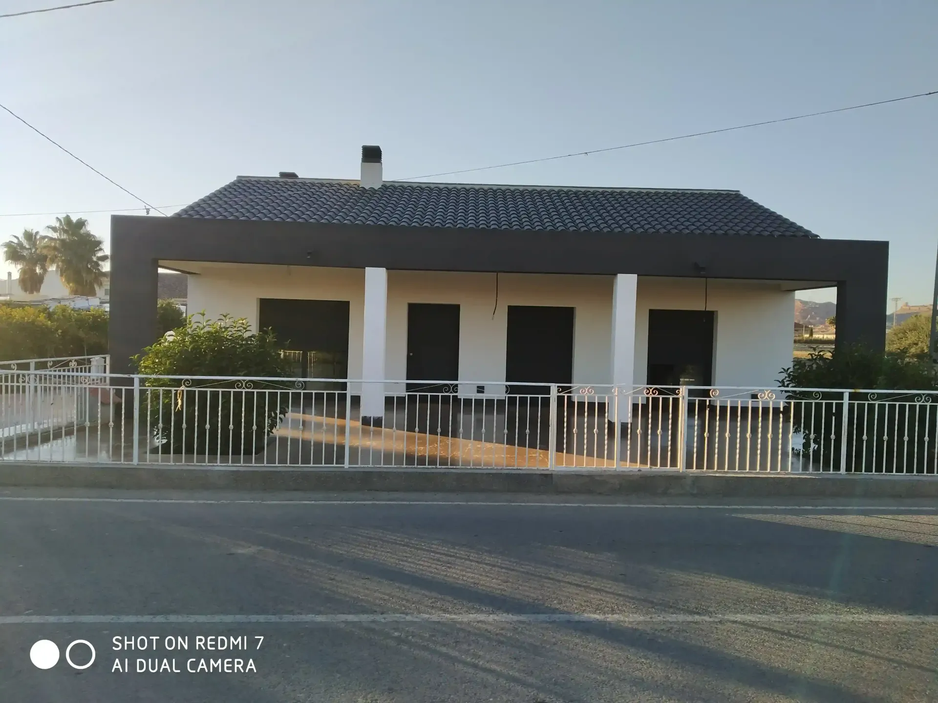 Casa de una sola planta con techo oscuro, columnas blancas y una valla blanca junto a una carretera.