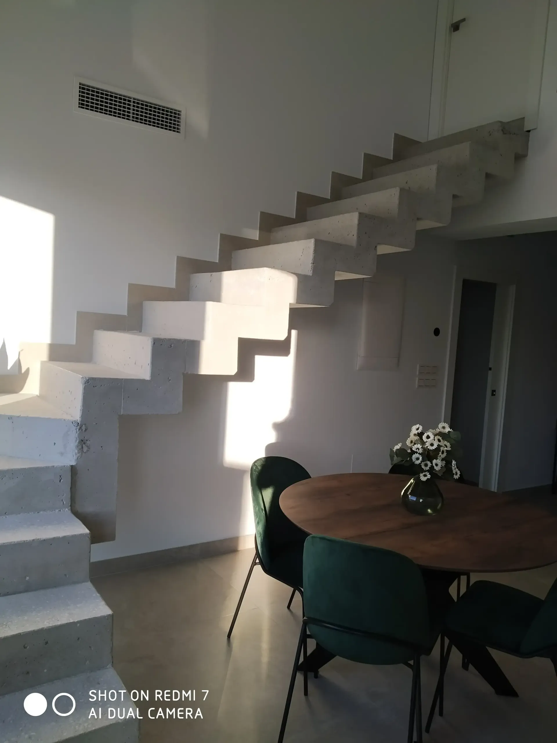 Interior moderno con escalera flotante, mesa redonda de madera y sillas bañadas por la luz del sol.
