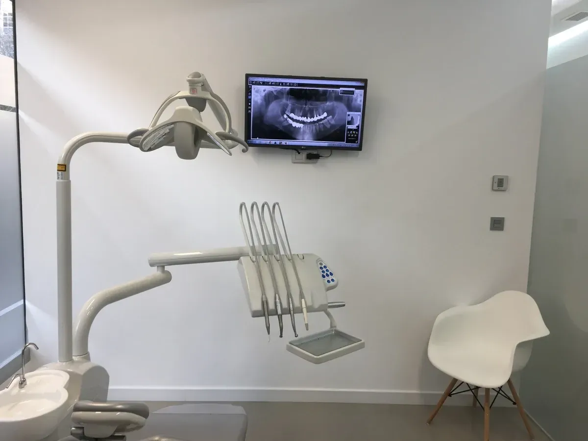 Sala de examen dental con silla, luz de techo, monitor y silla blanca contra una pared lisa.
