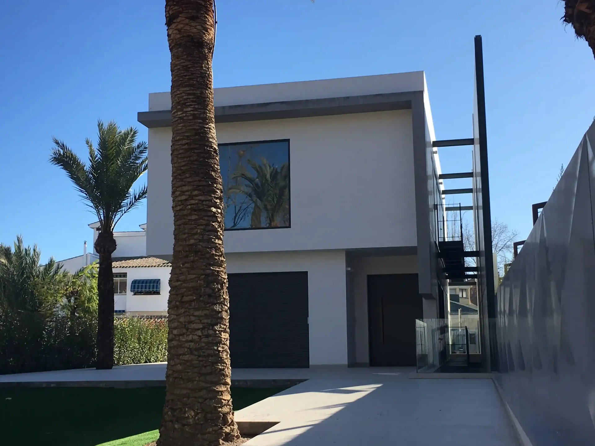 Casa moderna de dos plantas con paredes blancas, garaje oscuro, palmera y entrada vallada bajo un cielo azul despejado.