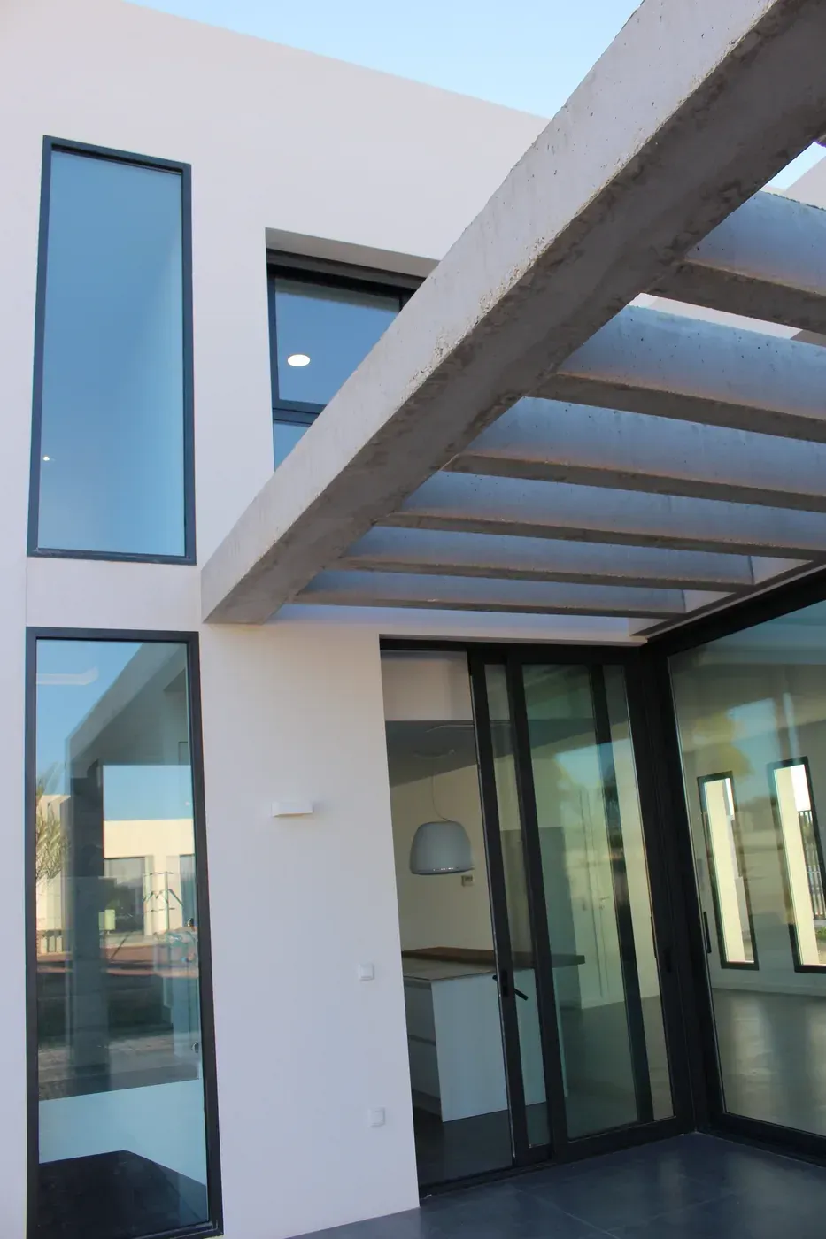 Entrada moderna al edificio con grandes ventanales, paredes blancas y una pérgola de cristal en la parte superior.