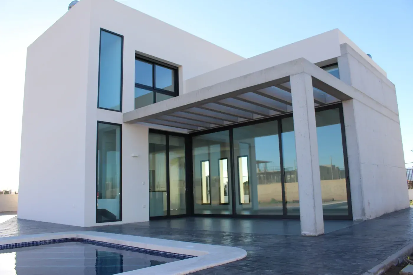 Casa moderna blanca con grandes ventanales y un patio cubierto junto a una piscina.