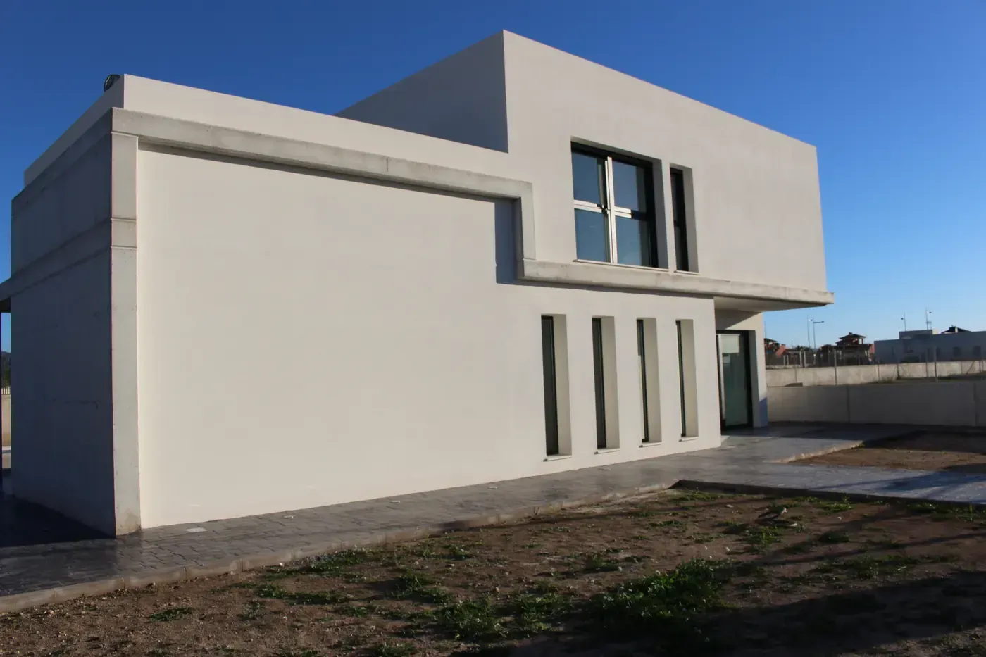 Moderna casa blanca de dos plantas con techo plano y grandes ventanales bajo un cielo azul despejado.