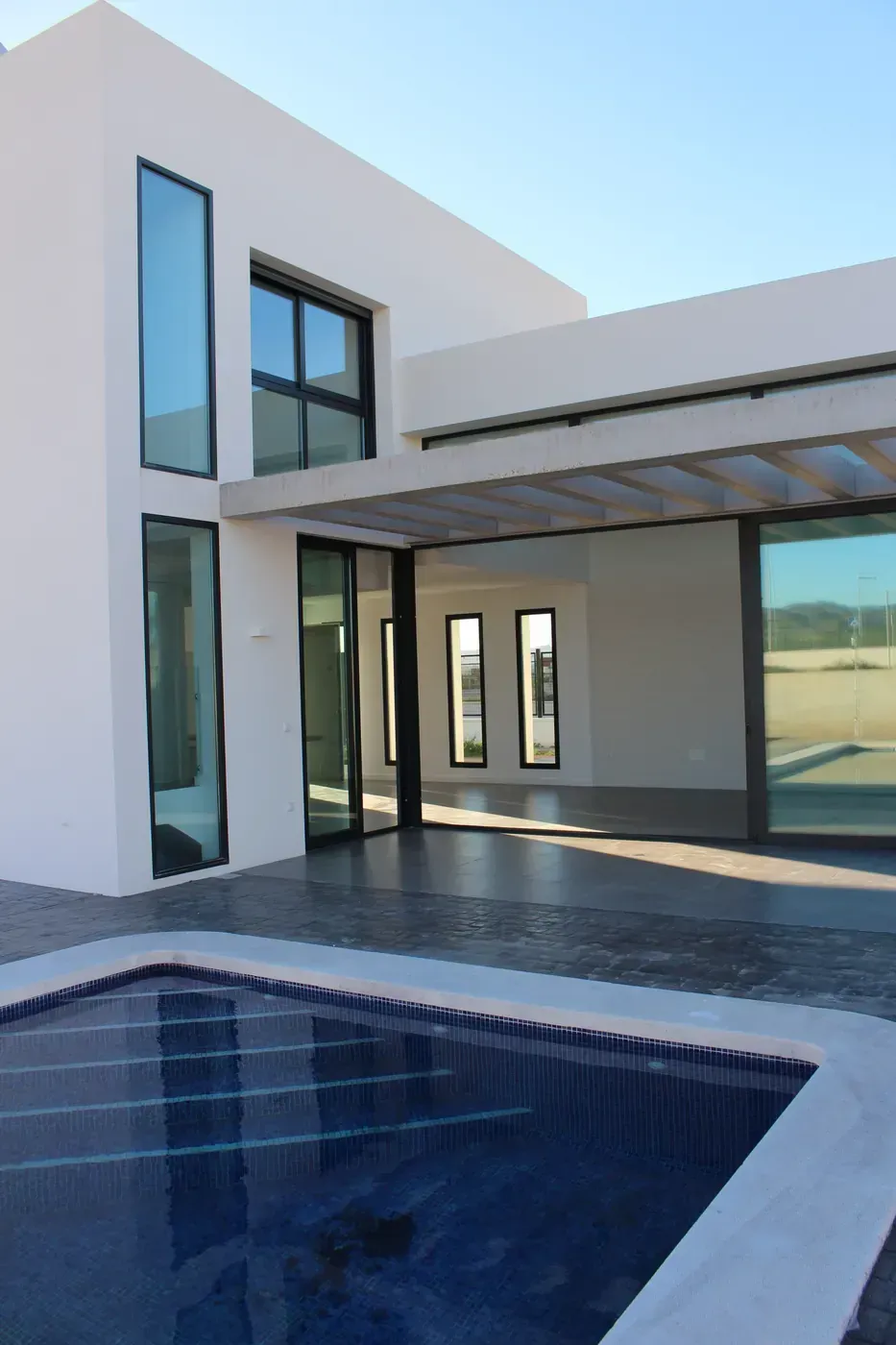 Casa blanca moderna con grandes ventanales y una piscina con azulejos azules en primer plano.