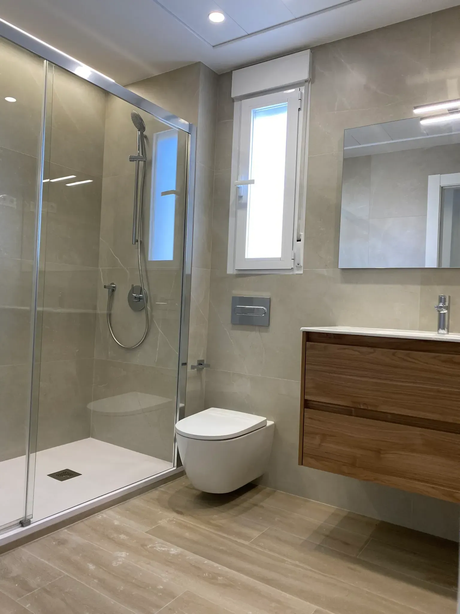 Baño moderno con ducha de cristal, inodoro suspendido, ventana y lavabo de madera.
