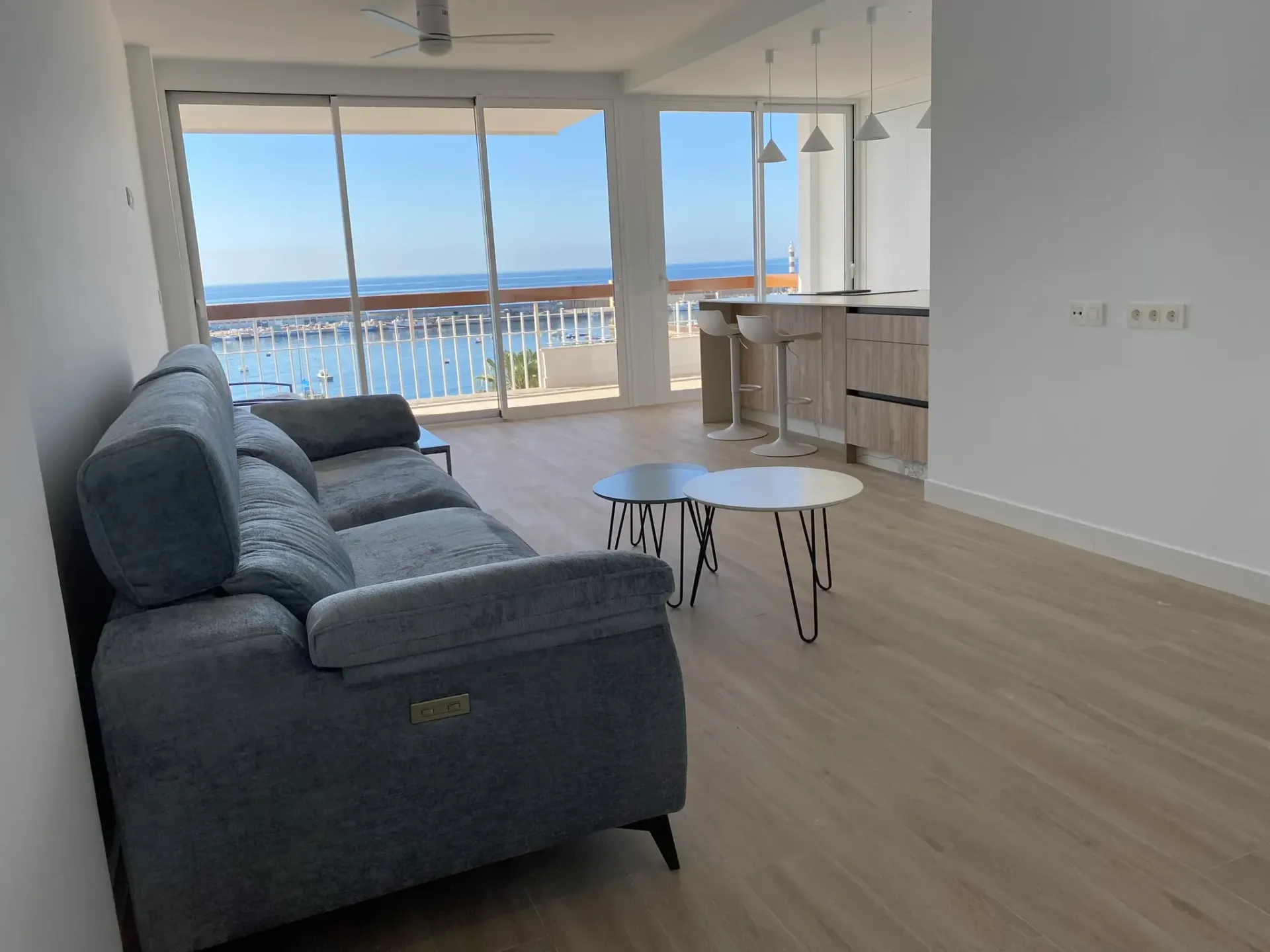 Sala de estar moderna con sofá azul, mesa de centro blanca y grandes ventanales con vistas al océano y al balcón.