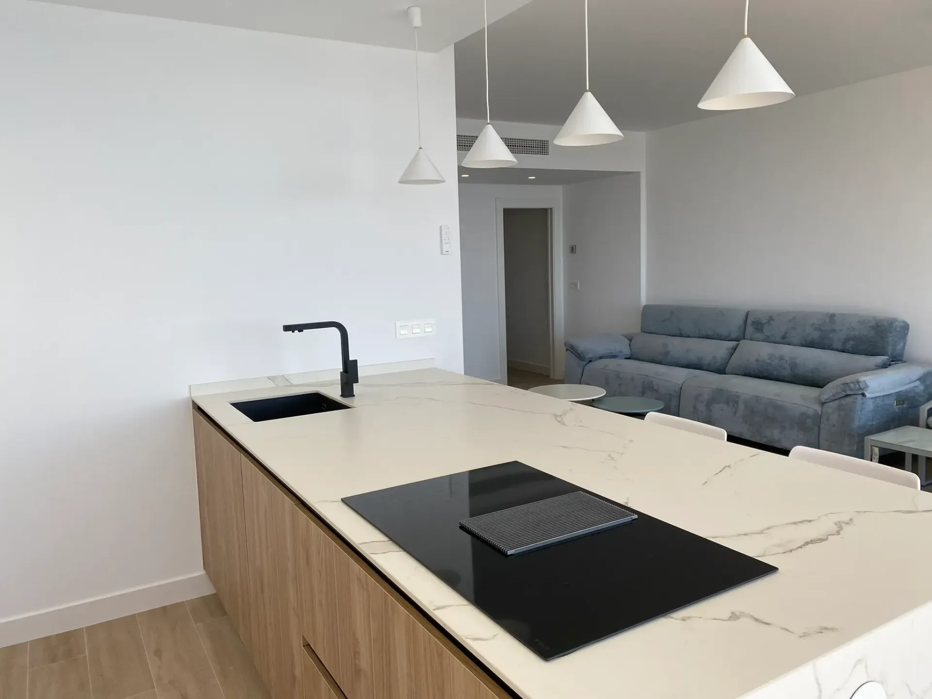 Isla de cocina moderna con encimera negra, lámparas colgantes y un sofá gris en una sala de estar abierta.