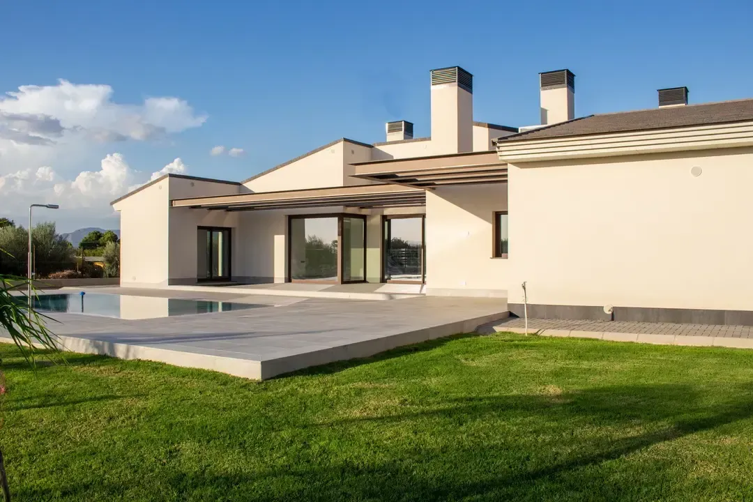 Casa moderna de color crema con patio y césped verde bajo un cielo azul.