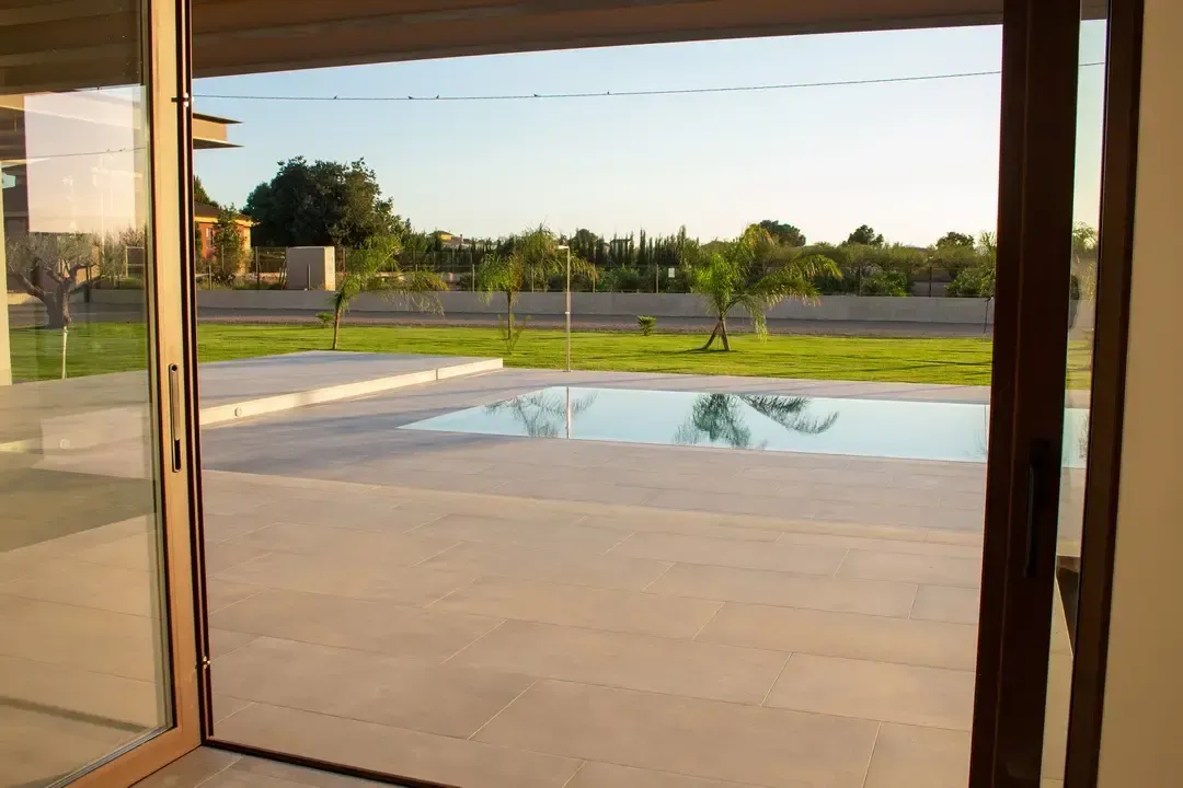 Vista del patio junto a la piscina a través de puertas abiertas hacia un soleado patio trasero y piscina.