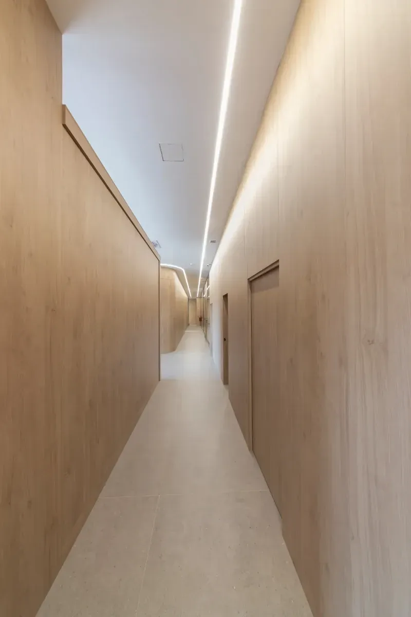 Pasillo estrecho y vacío con paredes de madera clara y una lámpara de techo fluorescente brillante.