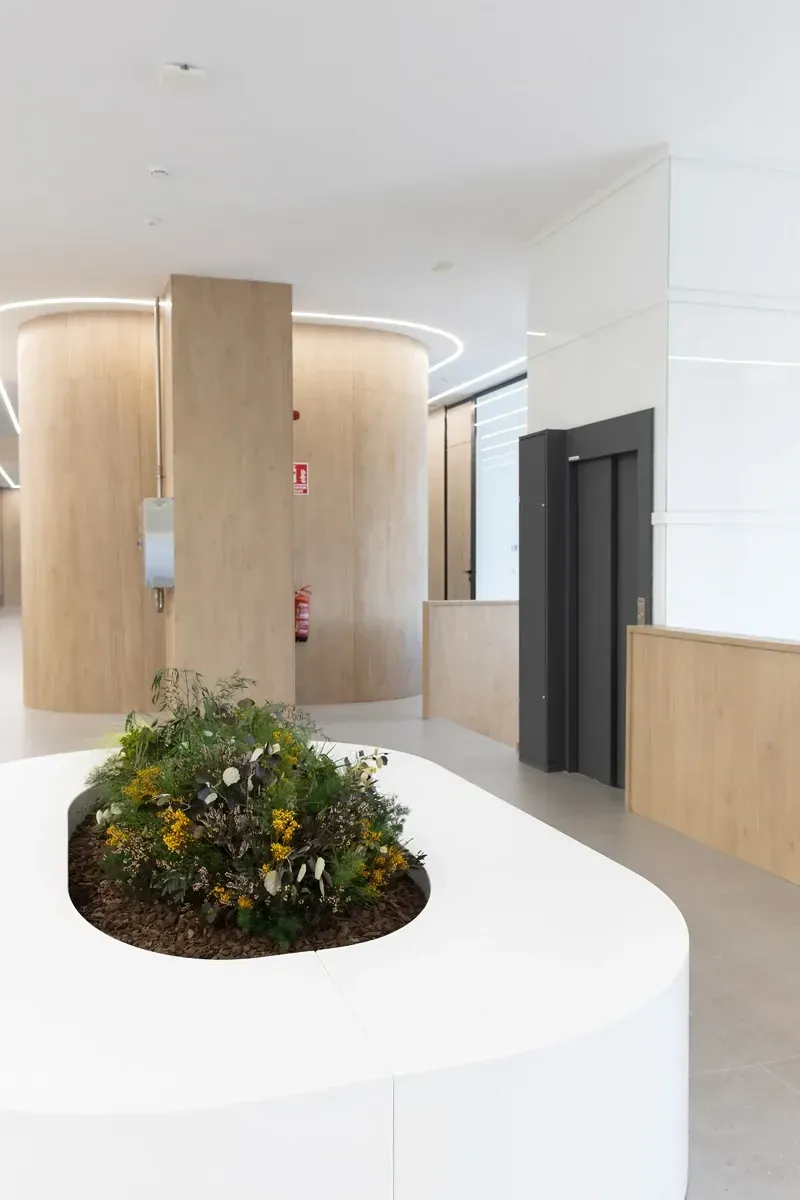 Vestíbulo moderno con jardinera curva blanca, vegetación, columnas de madera y puerta de ascensor.