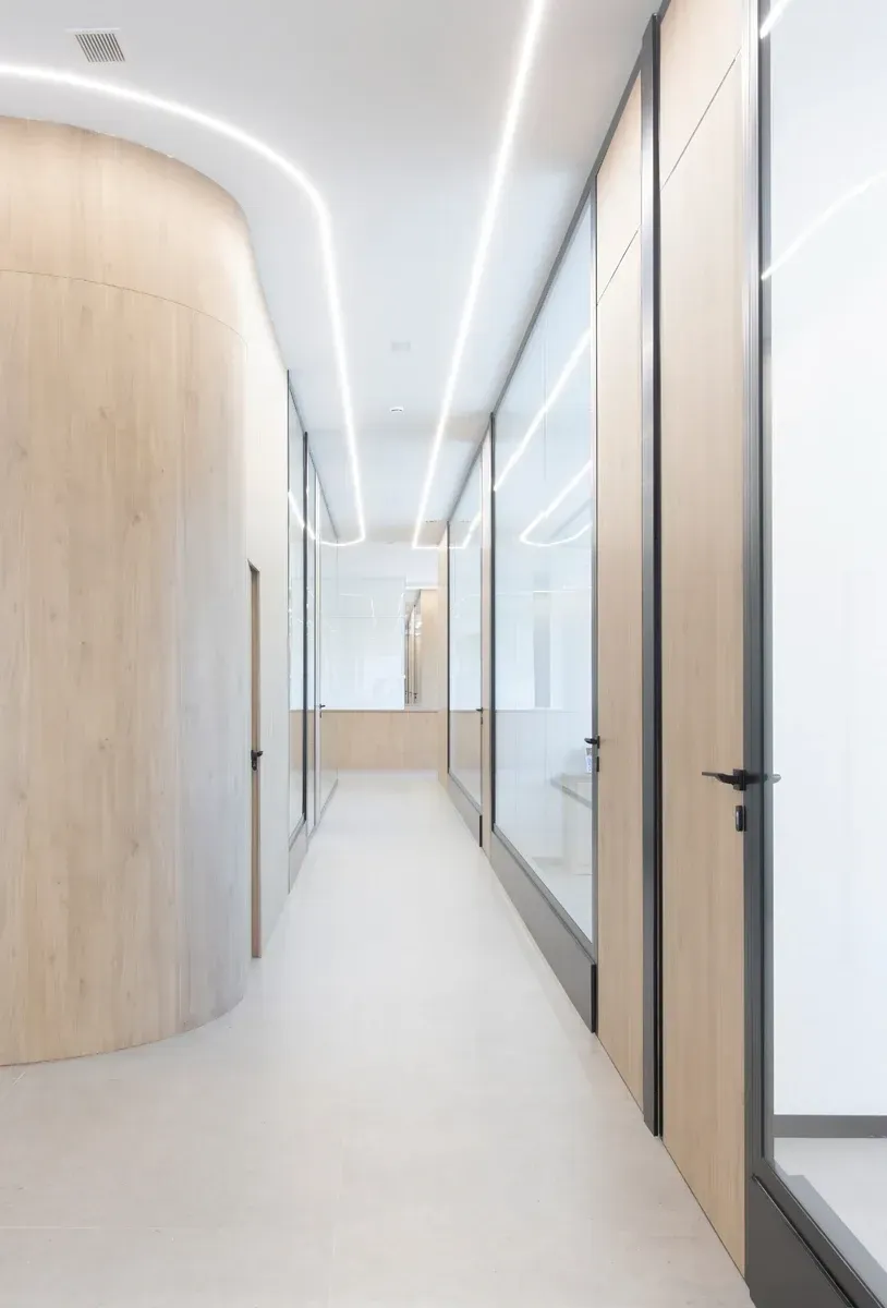Pasillo de oficina moderno con paredes de cristal, puertas de madera clara y luces de techo curvas.