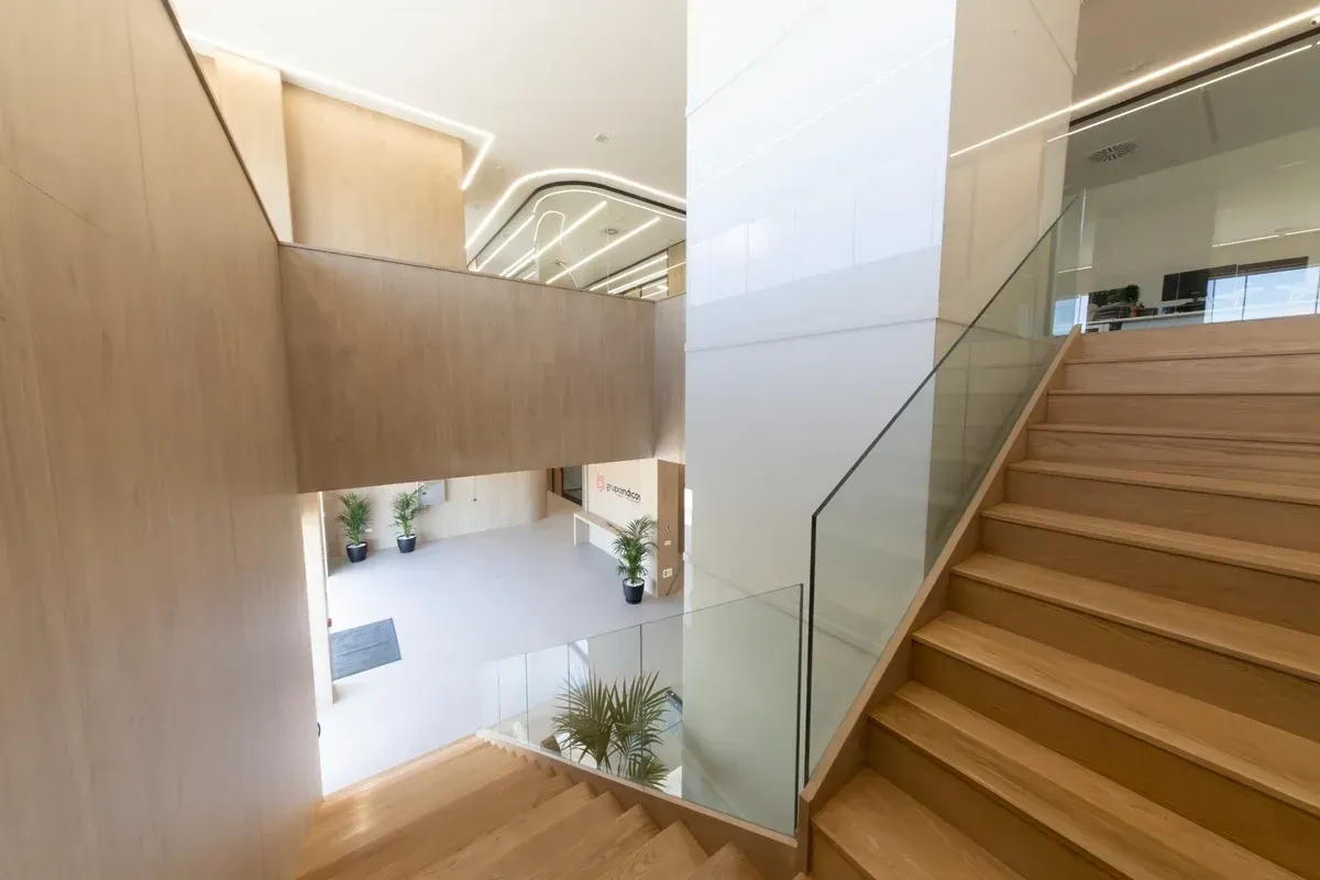 Escalera interior moderna con escalones de madera, barandilla de cristal y paredes de madera clara en un atrio abierto.