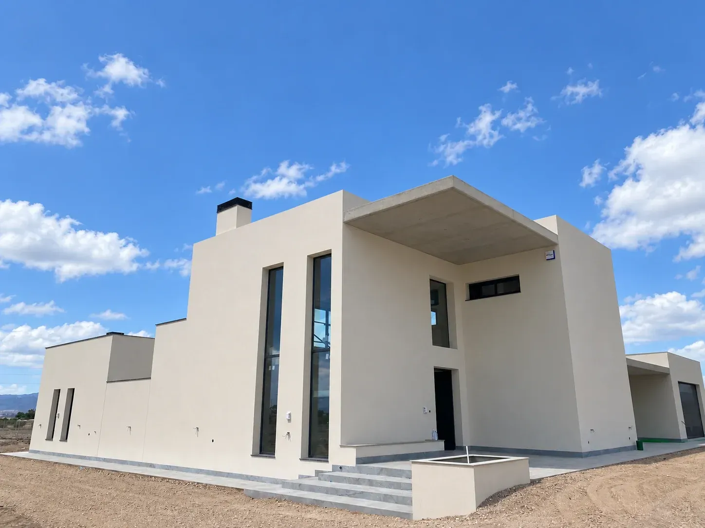 Casa moderna de color crema con diseño geométrico y un porche cubierto bajo un cielo azul.