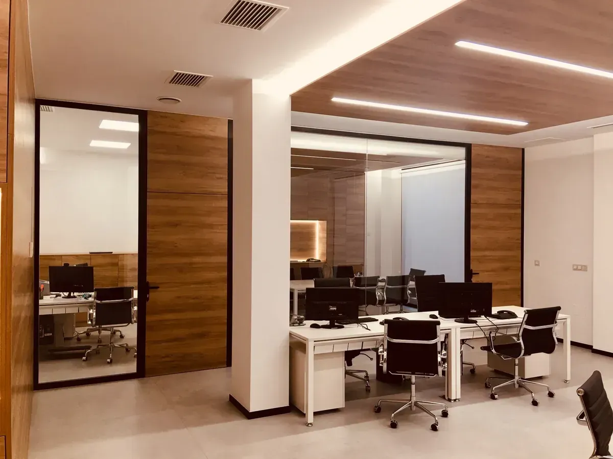 Oficina moderna con escritorios, monitores negros, sillas giratorias, paredes revestidas de madera y columnas blancas.