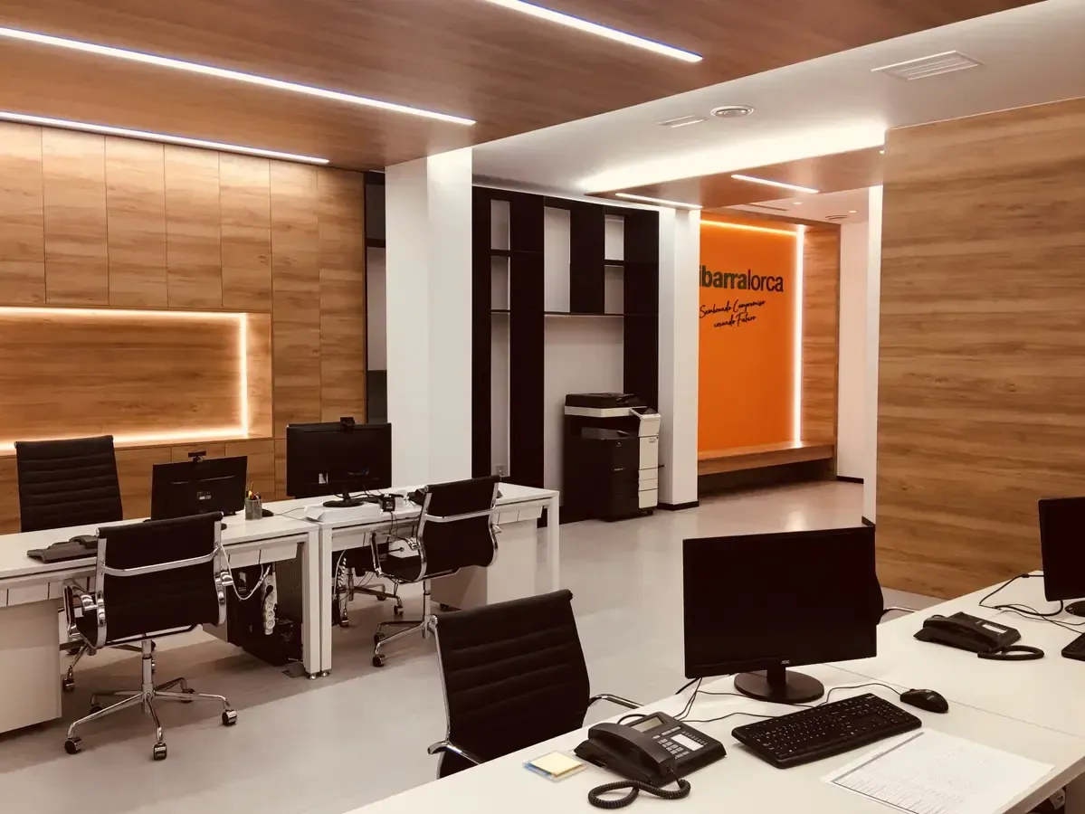 Interior de oficina moderno con escritorios, ordenadores, sillas negras y paredes revestidas de madera cálida.